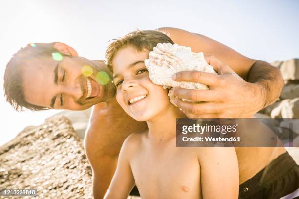 father and son having fun on the beach and listening to the ocean in a shell - sea shell stock-fotos und bilder