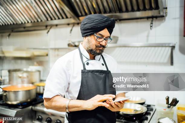 indian chef using smartphone in restaurant kitchen - kopfschmuck accessoires stock-fotos und bilder
