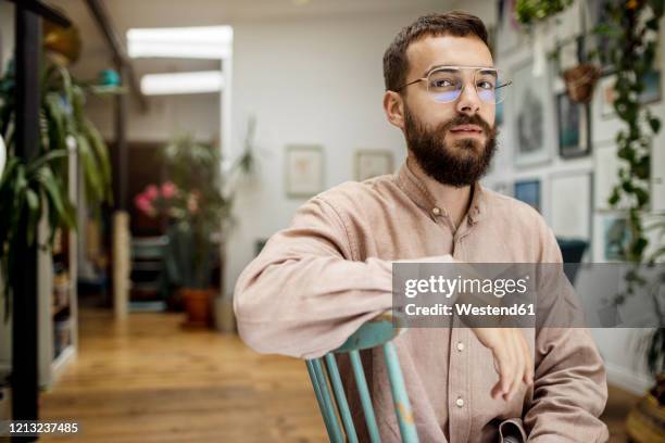 young man sitting on chair, looking at camera sceptically - suspicion stock-fotos und bilder