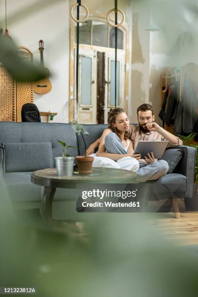 young couple sitting on couch at home, using digital tablet - couple tablet house stock-fotos und bilder