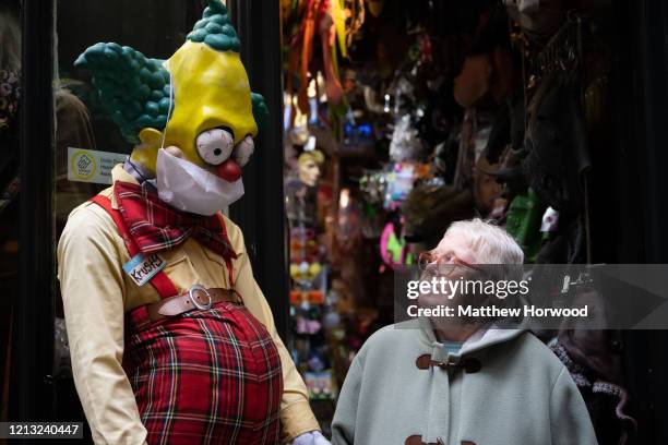 An elderly woman walks passed a Simpsons character in a surgical face mask outside a fancy dress shop on March 18, 2020 in Cardiff, United Kingdom....