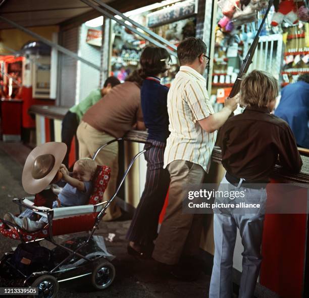 Ein Familienvater versucht auf einem Kirmesplatz sein Glück an einem Schießstand. Das Jüngste im Kinderwagen spielt derweil mit einer Puppe, die die...