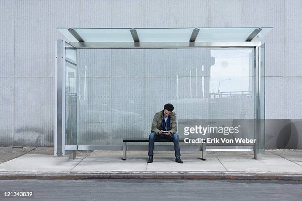 man sitting at glass bus stop with handheld device - banco asiento fotografías e imágenes de stock