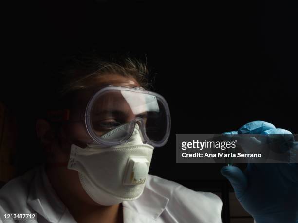 a female doctor or scientist wearing a mask, goggles and a lab coat analyzing a sample with protective gloves - low key imagens e fotografias de stock