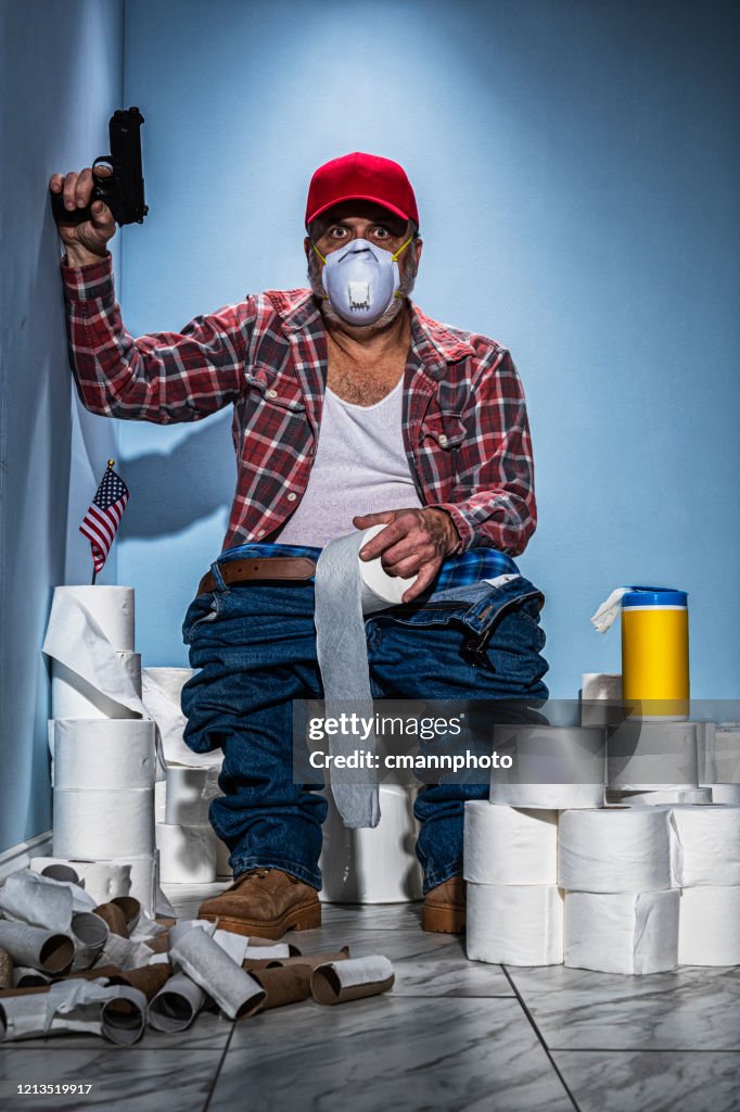 Man sitting on toilet guarding his stash of toilet paper with a handgun