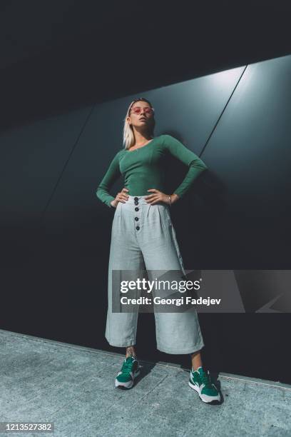 long haired fashion blonde, standing around the black wall, wearing a green cotton sweater, white wide pants, headband and sunglasses. bright sunlight flare falls on background. - groene schoen stockfoto's en -beelden