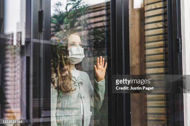 young girl looking through window with mask - quarantine stock pictures, royalty-free photos & images