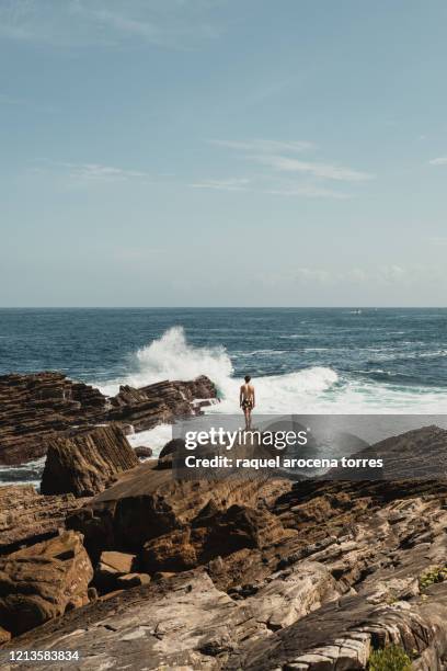 rear view of young white man in swimsuit stand up on the rocks by the sea - hondarribia stock-fotos und bilder