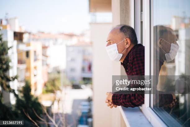 senior mann in medizinischer maske auf fenster atmen frische luft aus quarantäne - quarantäne stock-fotos und bilder