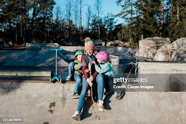 mom and her kids laughing having fun and playing at skatepark - mother and daughter riding on skateboard in park stock pictures, royalty-free photos & images