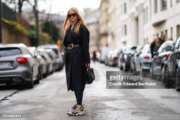 Charlotte Groeneveld wears sunglasses, earrings, a turtleneck top, a black long coat, a leather and golden belt, leggings, a leather bag, Balenciaga...
