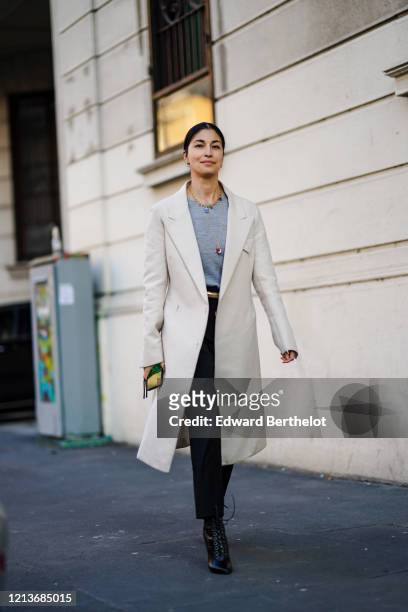 Caroline Issa wears earrings, necklaces, a heather blue-grey top, a white coat, a gold-tone belt, black pants, black pointy heeled boots with...