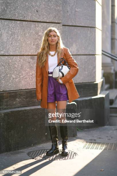 Emili Sindlev wears earrings, a necklace, a white top, a white belt, a tan-color leather long jacket, purple leather shorts, black knee-high boots, a...