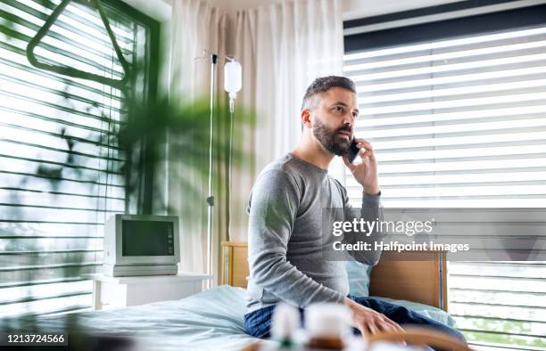 ill mature man patient sitting on bed in hospital, using smartphone. - adult male hospital bed stock pictures, royalty-free photos & images