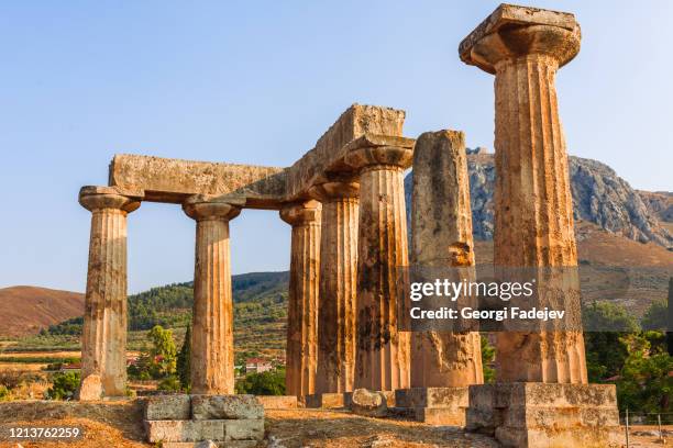 ruins of temple of apollo in corinth greece standing up on a hill with remenants of rock walls scattered about under a bright sun with mountians and a blue sky behind - templo de apolo corinto imagens e fotografias de stock