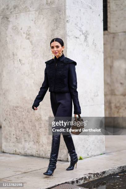Nathalie Fanj wears large earrings, a black jacket with embroidery, a bronze color bag with embroidery, a skirt, black leather pointy high heels...