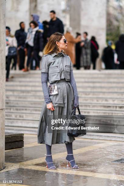 Natalia Ostrofsky wears sunglasses, a gray turtleneck top, a silver shiny bag, a gray pleated long jacket/dress, with pockets on the upper part,...