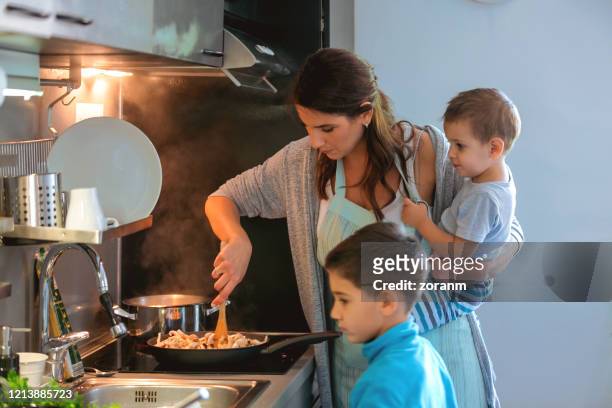 madre sosteniendo a un niño pequeño y cocinando, hijo mayor de pie - madre ama de casa fotografías e imágenes de stock