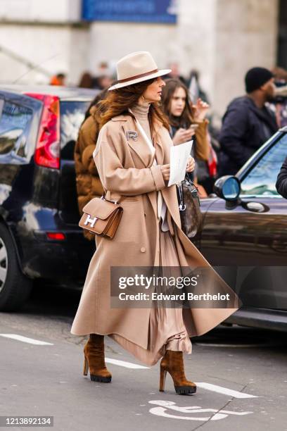 Guest wears a hat, a beige long coat, a Hermes brown leather bag, a flowing skirt, a turtleneck pullover, brown suede high heels shoes, outside Elie...