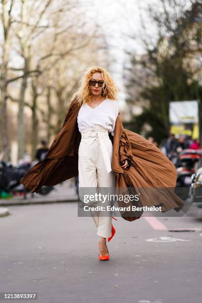 Elina Halimi wears sunglasses, a white v-neck t-shirt, a brown flowing long coat, white pants, neon orange pointy high heeled shoes, outside Elie...