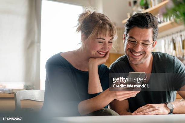 happy couple sitting at table in kitchen using smartphone - mid adult men stock pictures, royalty-free photos & images