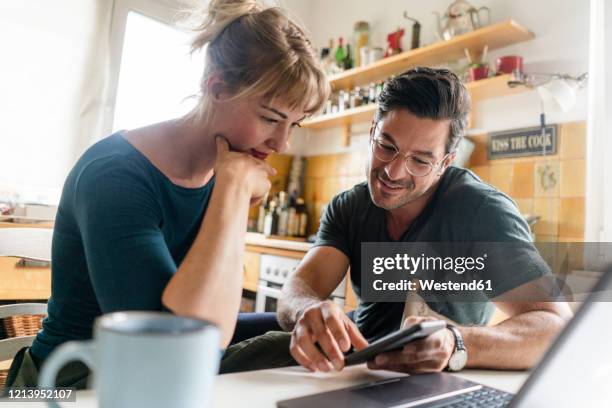 couple sitting at table in kitchen using smartphone - gedeelde mobiliteit stockfoto's en -beelden