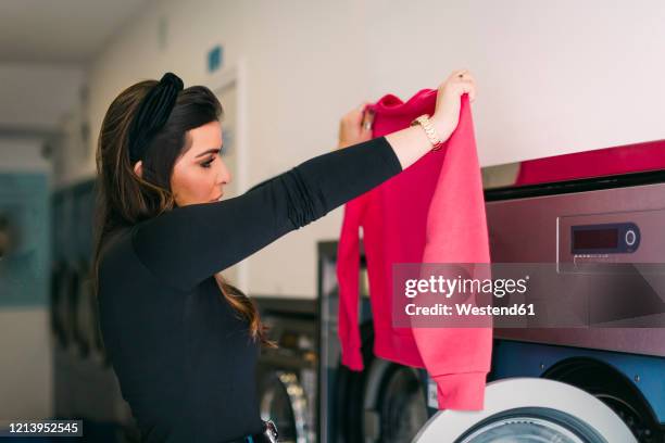 young woman doing the laundry in a launderette looking at pink sweatshirt - laundry woman stock pictures, royalty-free photos & images