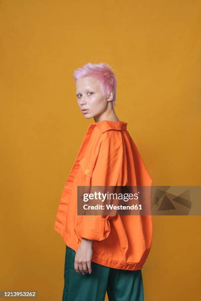 portrait of young woman with short pink hair wearing orange jacket in front of yellow background - punk stock pictures, royalty-free photos & images