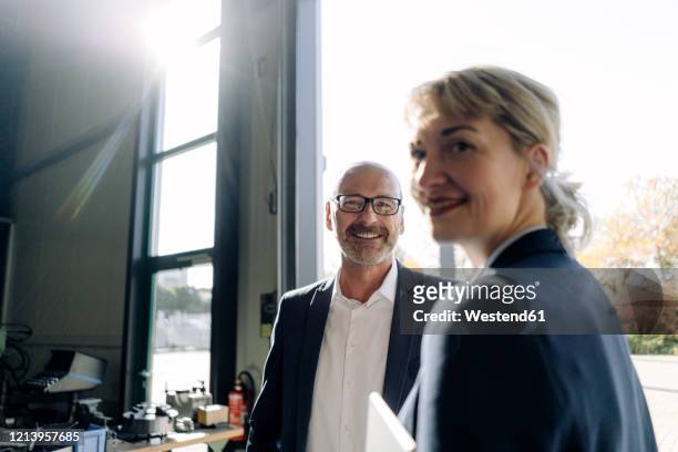 portrait of smiling businessman and businesswoman at the window in a factory - geschäftsleben stock-fotos und bilder