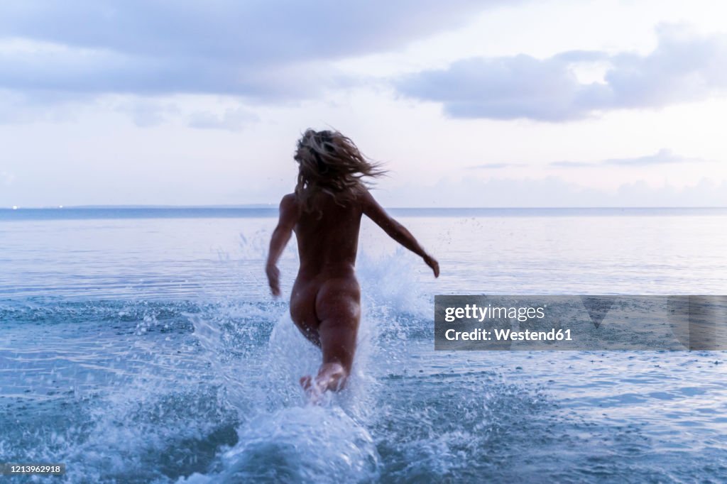 Back view of young woman running into the sea at sunset