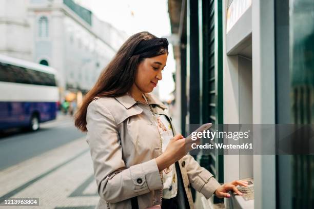 woman using atm machine and mobile phone - dab photos et images de collection