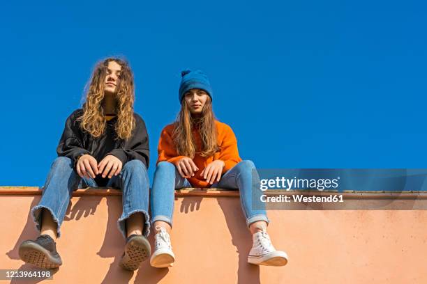 portrait of teenage girls sitting on a wall against blue sky - vista de ángulo bajo fotografías e imágenes de stock