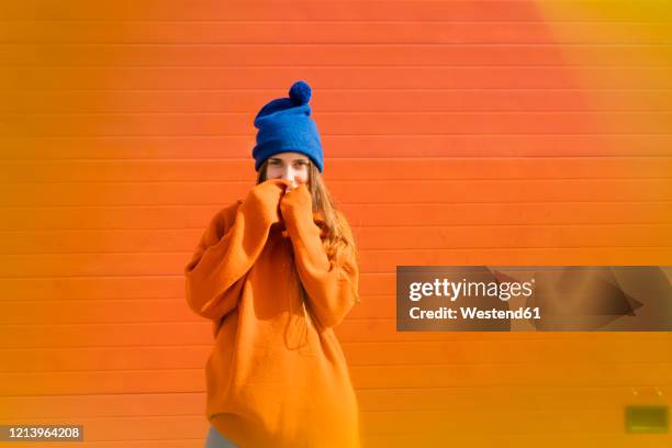portrait of teenage girl weraing blue woolly hat and orange sweater in front of orange background - mão na boca imagens e fotografias de stock