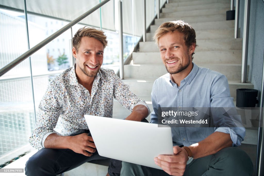 Portrait of two smiling young businessmen sitting on stairs with laptop