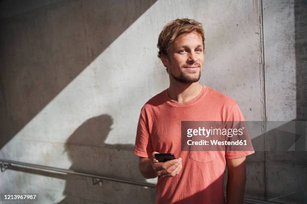 portrait of man wearing red t-shirt holding cell phone at concrete wall - mid volwassen mannen stockfoto's en -beelden