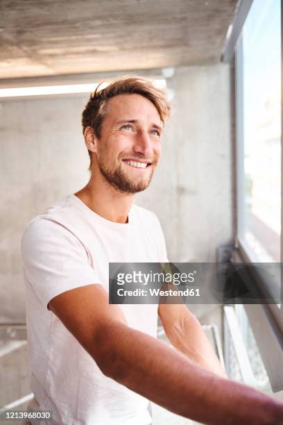 portrait of smiling young man wearing white t-shirt looking out of window - seitenblick stock-fotos und bilder