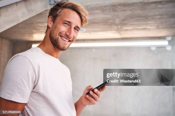 portrait of attactive young man wearing white t-shirt holding smartphone - auslachen blick in die kamera stock-fotos und bilder