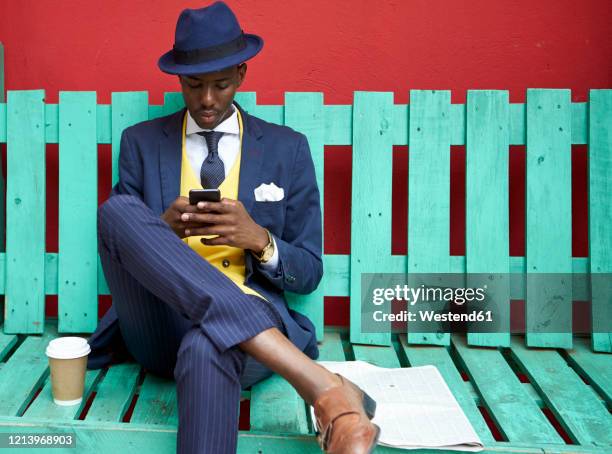 young businessman wearing old-fashioned suit and hat sitting on a green bench checking his phone - multi coloured suit stock pictures, royalty-free photos & images