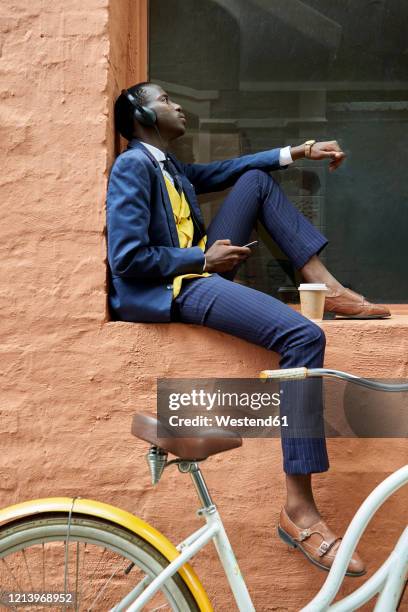 stylish young businessman with bicycle wearing old-fashioned suit sitting on a the ledge of a window listening to music on his headphones - menswear bildbanksfoton och bilder