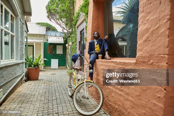 stylish young businessman with bicycle wearing old-fashioned suit sitting on a the ledge of a window listening to music on his headphones - businessman in black suit photos et images de collection