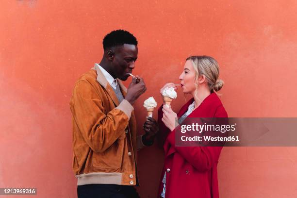 young couple standing at an orange wall eating ice cream - red couple stock-fotos und bilder