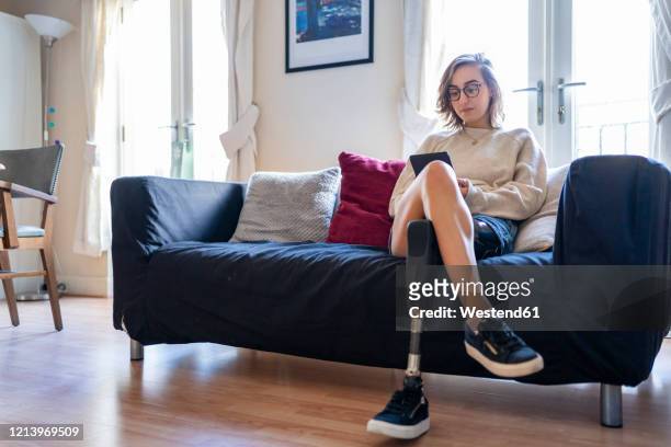 young woman with leg prosthesis sitting on couch at home reading e-book - amputee woman imagens e fotografias de stock