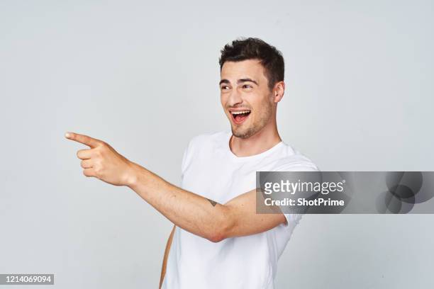 joyful young man in a white t-shirt with his finger pointing to the side on a blank gray background. - portrait of business man looking surprised stock-fotos und bilder
