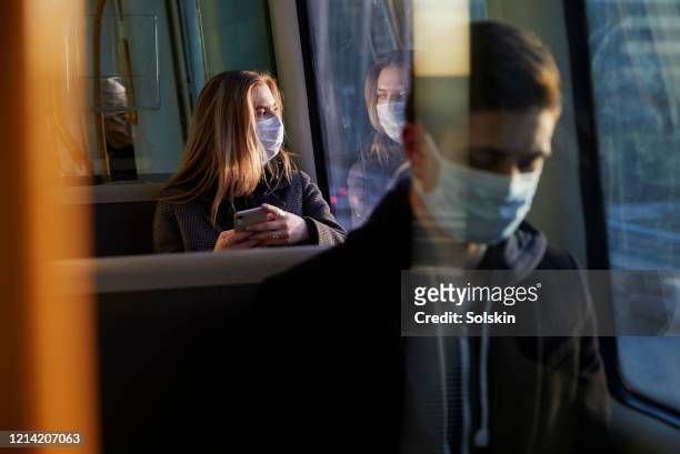 young woman sitting in train wearing protective mask, using smartphone - hauptverkehrszeit stock-fotos und bilder