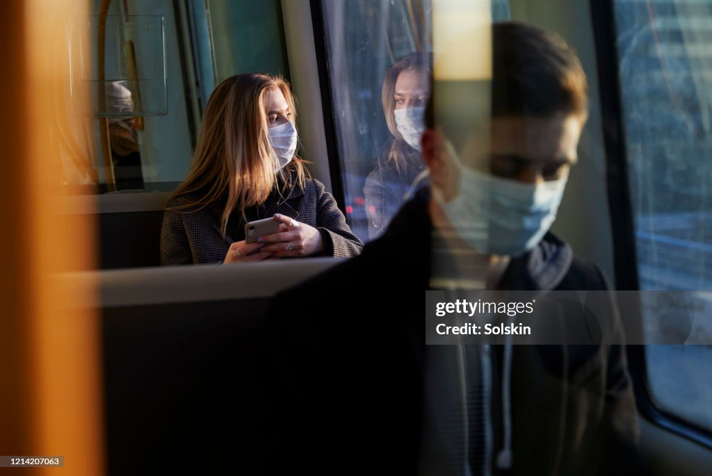 Young woman sitting in train wearing protective mask, using smartphone
