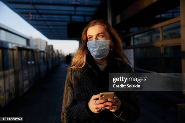 young woman standing on train station wearing protective mask, using phone - uncertainty pandemic stock pictures, royalty-free photos & images