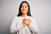 Young beautiful asian woman wearing casual sweater standing over white background smiling with hands on chest with closed eyes and grateful gesture on face. Health concept.