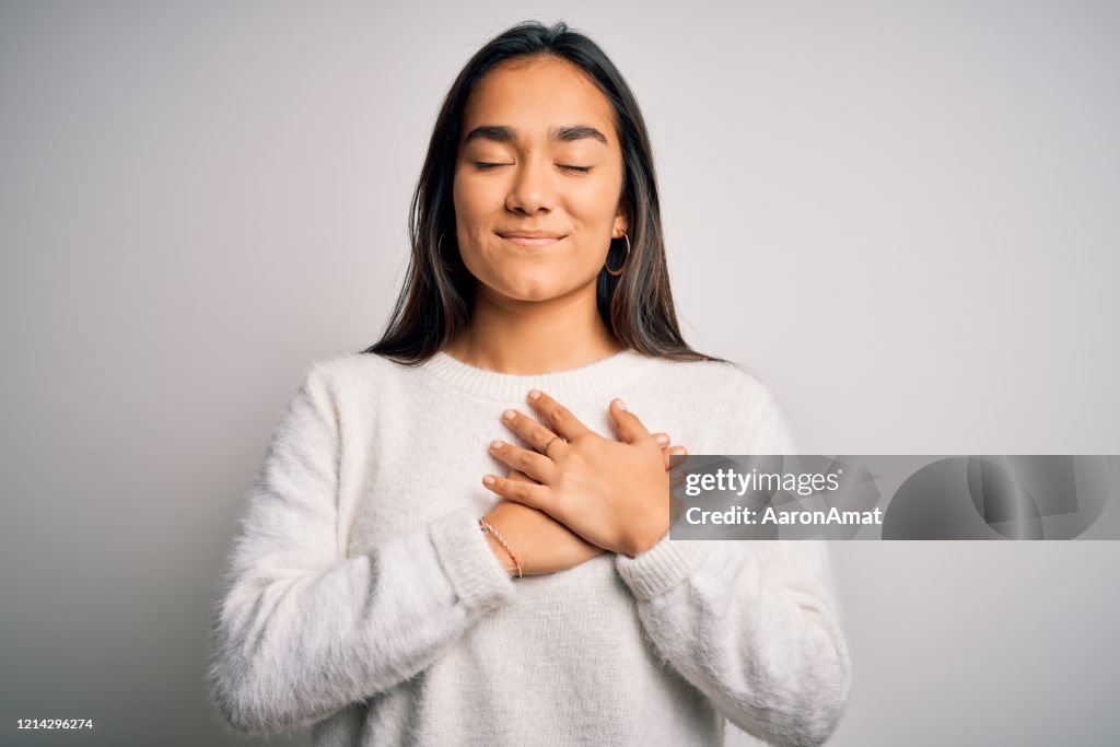 Young beautiful asian woman wearing casual sweater standing over white background smiling with hands on chest with closed eyes and grateful gesture on face. Health concept.