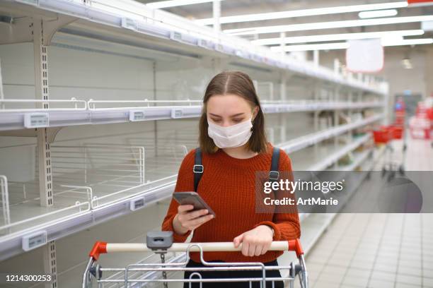 young woman standing near empty shelf in a supermarket and using smartphone - supermarket mask stock pictures, royalty-free photos & images