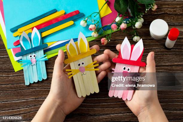 Child is holding Easter bunny toy gift stics puppets on wooden table. Handmade. Project of children's creativity, handicrafts, crafts for kids.